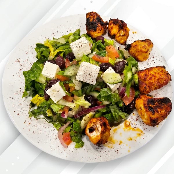 A white plate with a fresh Greek salad topped with feta cheese and grilled chicken pieces.