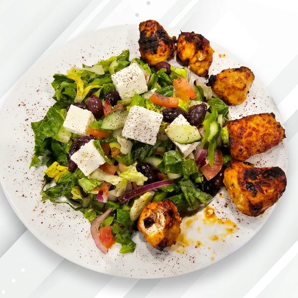 A white plate with a fresh Greek salad and grilled chicken pieces, sprinkled with spices.